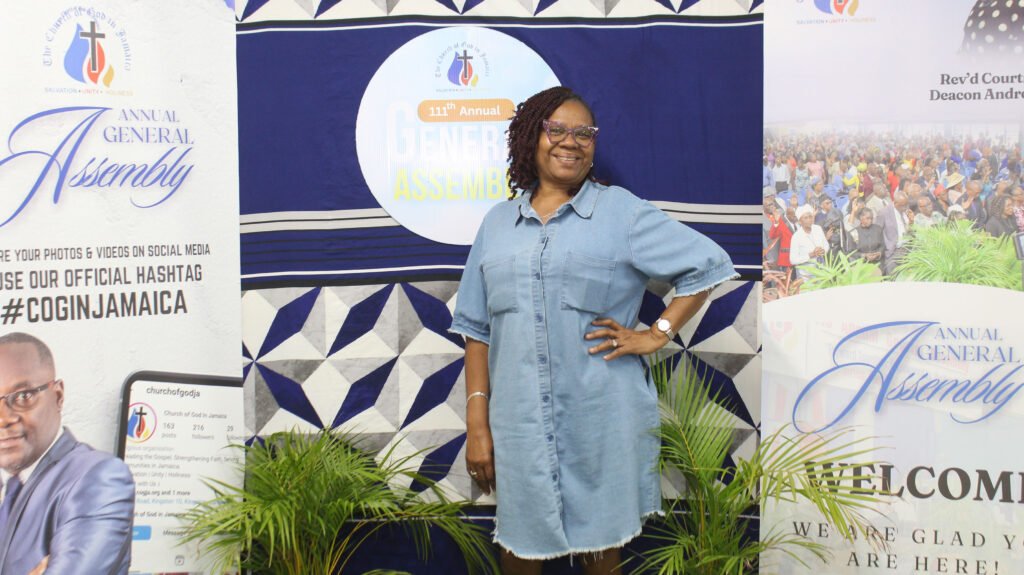 A woman in a denim dress smiles while posing for a photo at an event backdrop featuring the text 'Annual General Assembly'.