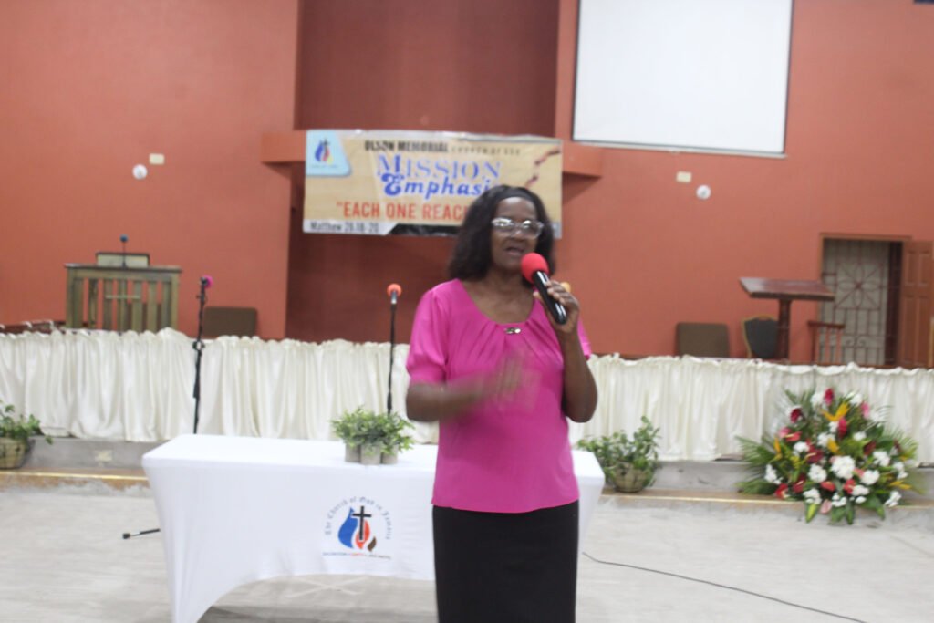 A woman in a pink shirt speaks at a church event, holding a microphone, with a white table in front adorned with plants.