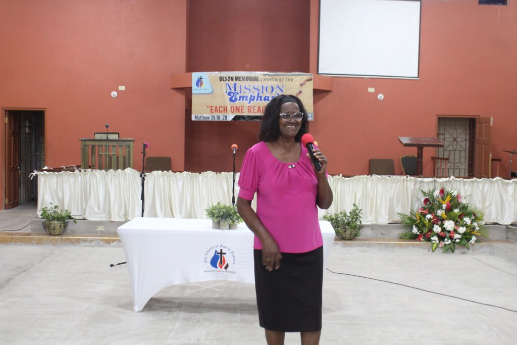 A woman in a pink top speaks into a microphone while standing in front of a table and a church backdrop, with decorations and a screen visible in the background.