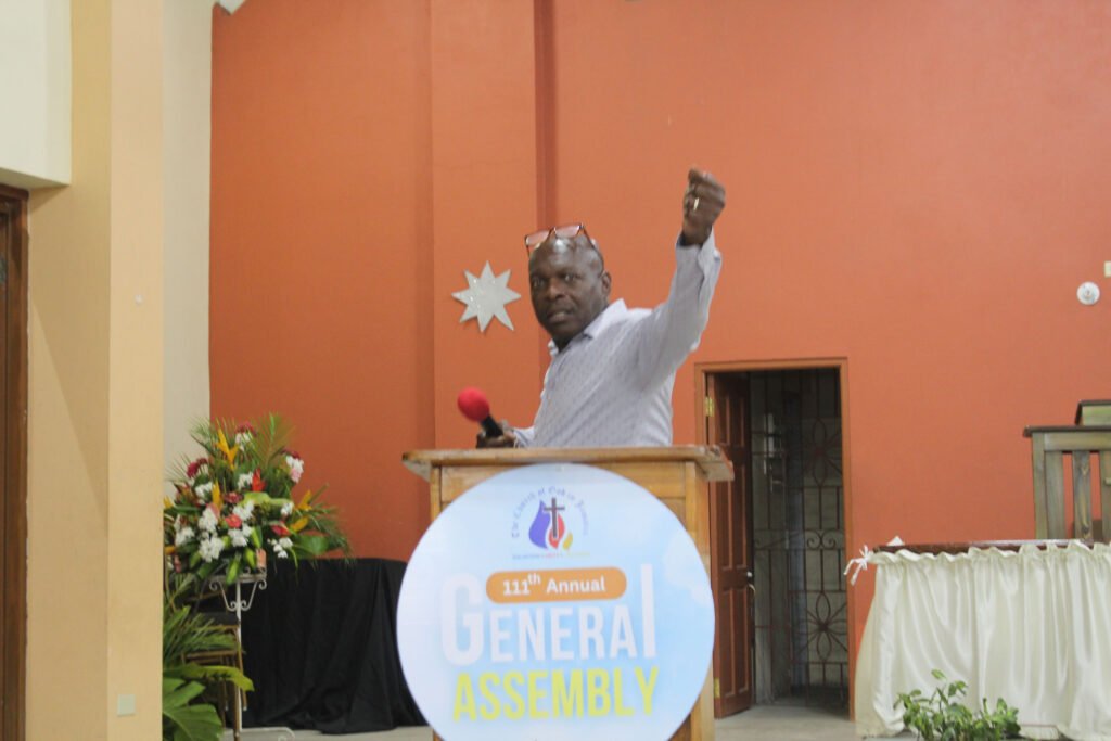 A man speaking passionately at a podium during a General Assembly event, with a colorful background and a floral arrangement nearby.