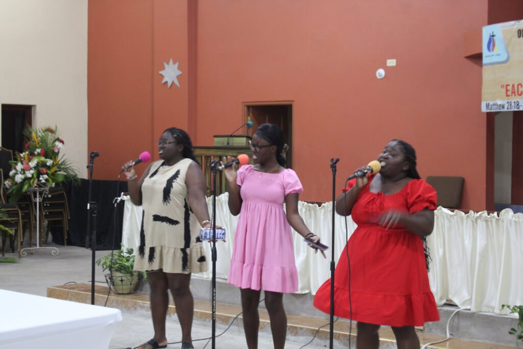 Three women singing at a microphone in a church setting, wearing colorful dresses. The interior features a flower arrangement and a banner with a religious message.