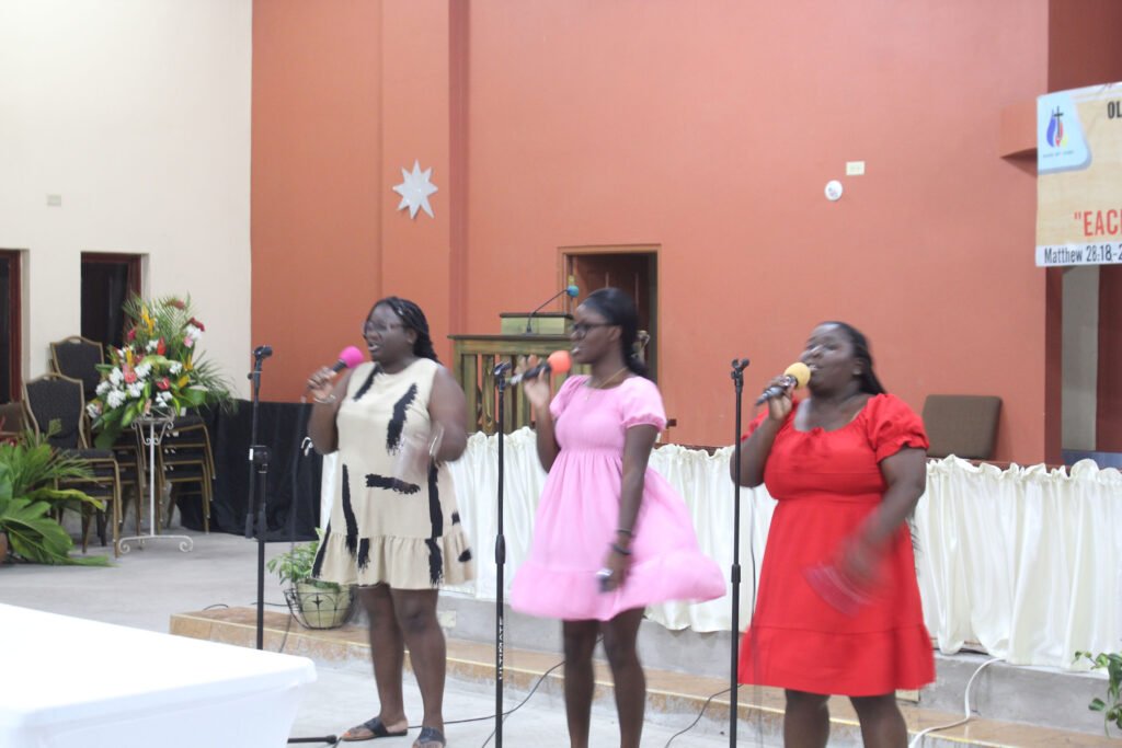 Three women singing into microphones during a performance in a church setting, with colorful dresses and a warm background.
