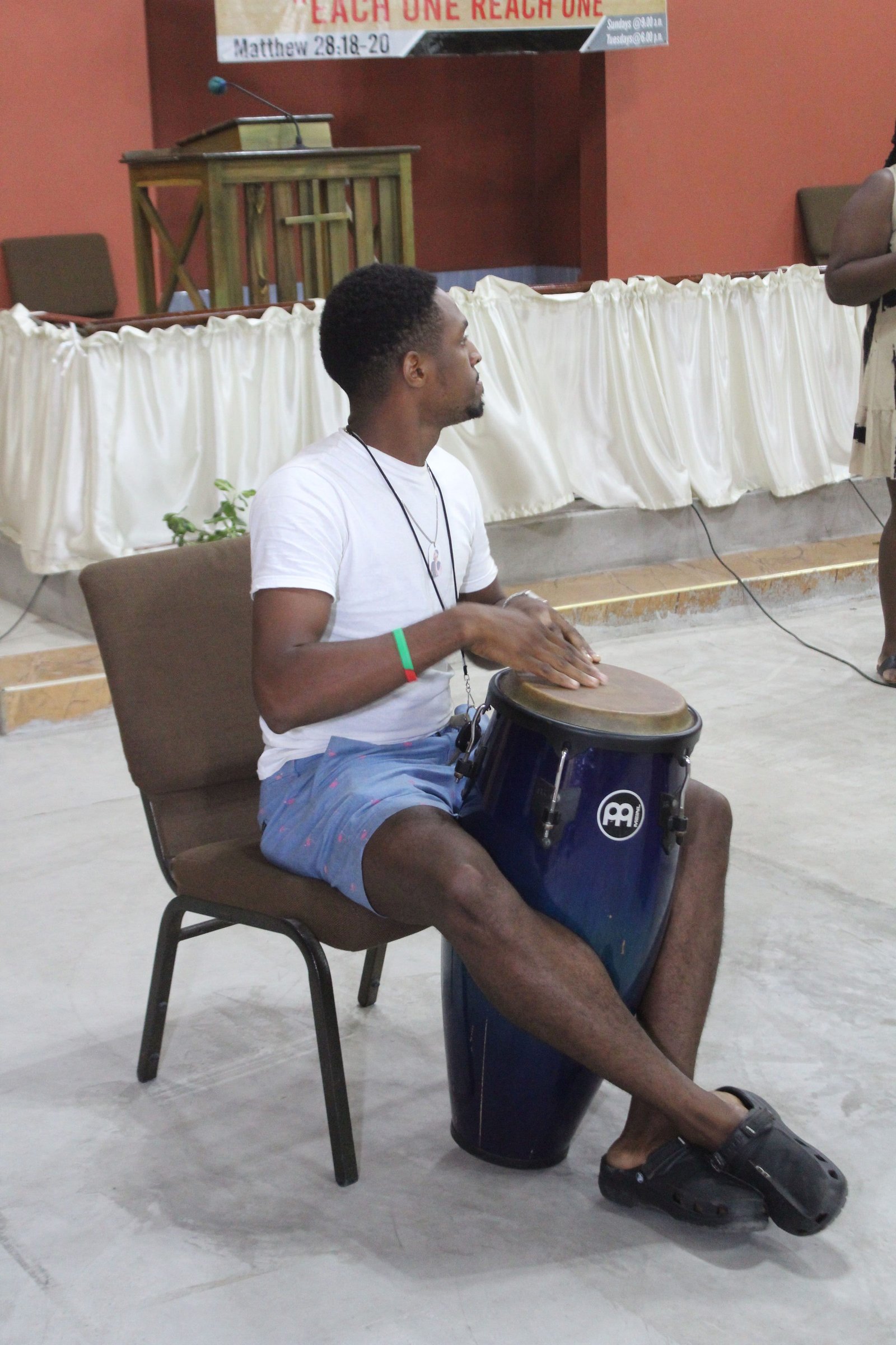 A person sitting on a chair, playing a conga drum with passion, wearing casual shorts and a white shirt in a spacious indoor setting.