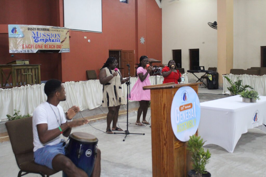 A group of performers singing on stage during a church event, with a drummer playing a conga in the foreground. Decorated backdrop featuring a mission emphasis banner and a general assembly sign.