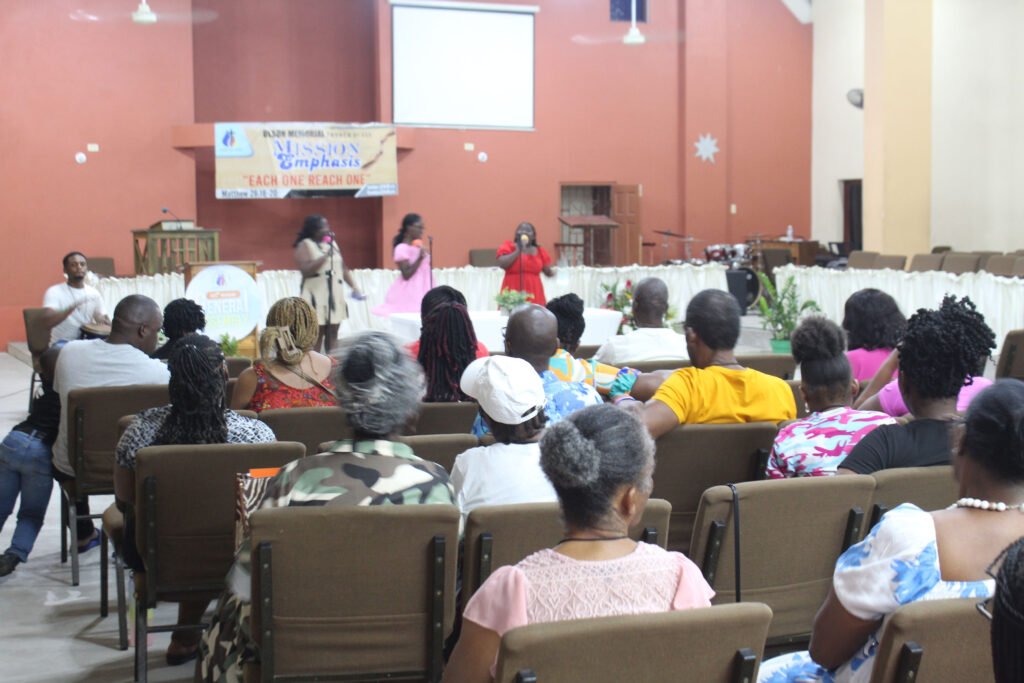 A gathering in a church setting with attendees sitting on chairs, listening to speakers on stage. Decorative flowers are present on the tables, and a banner reading 'MISSION EMPHASIS' is displayed in the background.