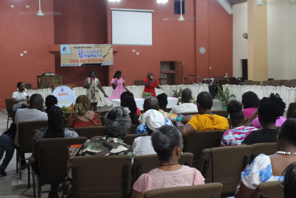 Indoor event with a presentation taking place on stage, an audience sitting in chairs, and decorations in the background.