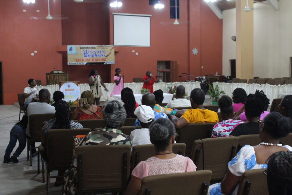Audience seated in a church during a missionary event, with speakers on stage and a banner in the background emphasizing the theme 'Each One Reach One'.