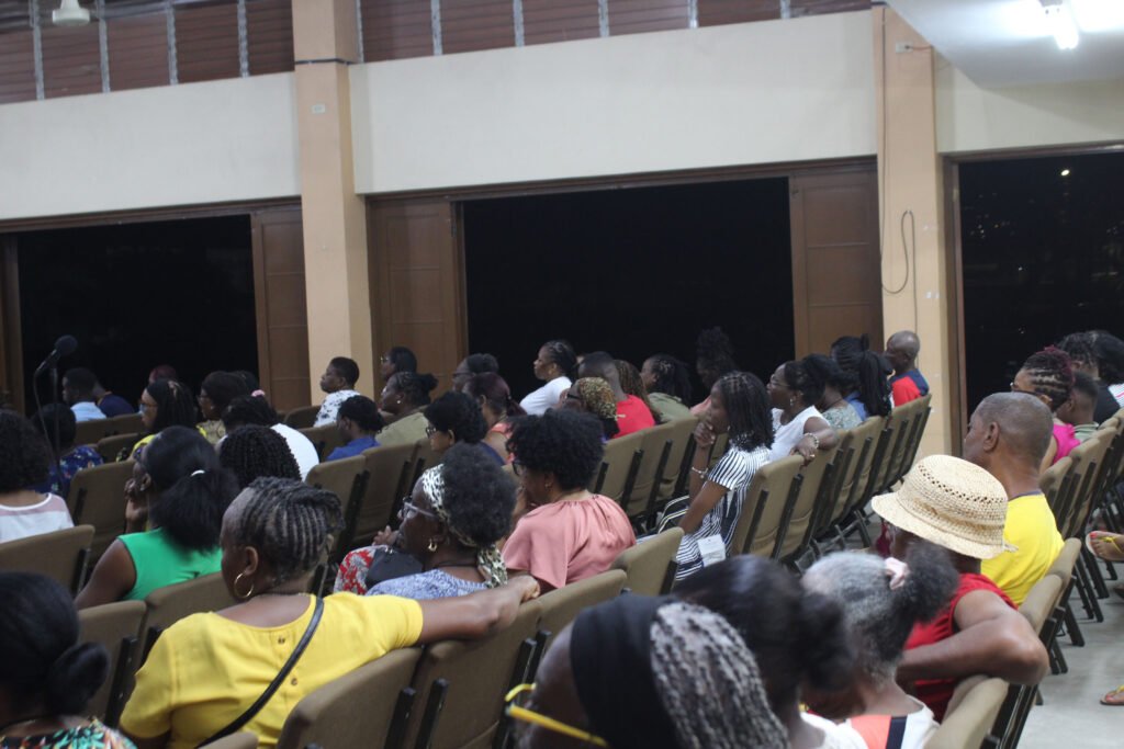 A crowd of diverse people sitting in an indoor venue during an event, with some attendees wearing colorful clothing and others with hats, focused on the front.
