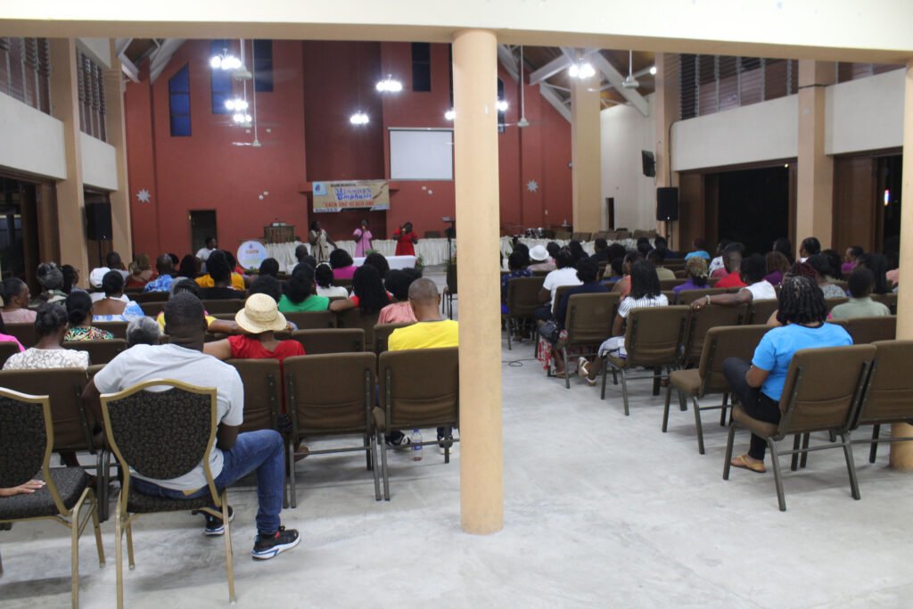 A gathering of people sitting in a hall, facing a stage where individuals are performing. The audience includes men and women of various ages, dressed in colorful clothing, with some wearing hats. The interior has high ceilings and decorative elements.