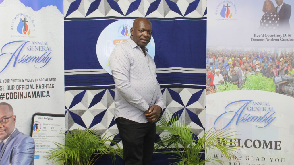 A man in a light-colored shirt stands in front of a backdrop featuring promotional banners for the Annual General Assembly, including information and hashtags for social media sharing.