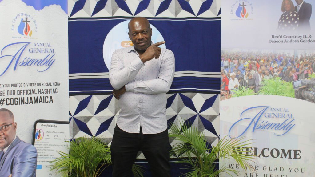 A man posing confidently with one hand on his hip and the other pointing upwards, set against a backdrop featuring banners for an Annual General Assembly, with details promoting social media engagement and welcoming attendees.
