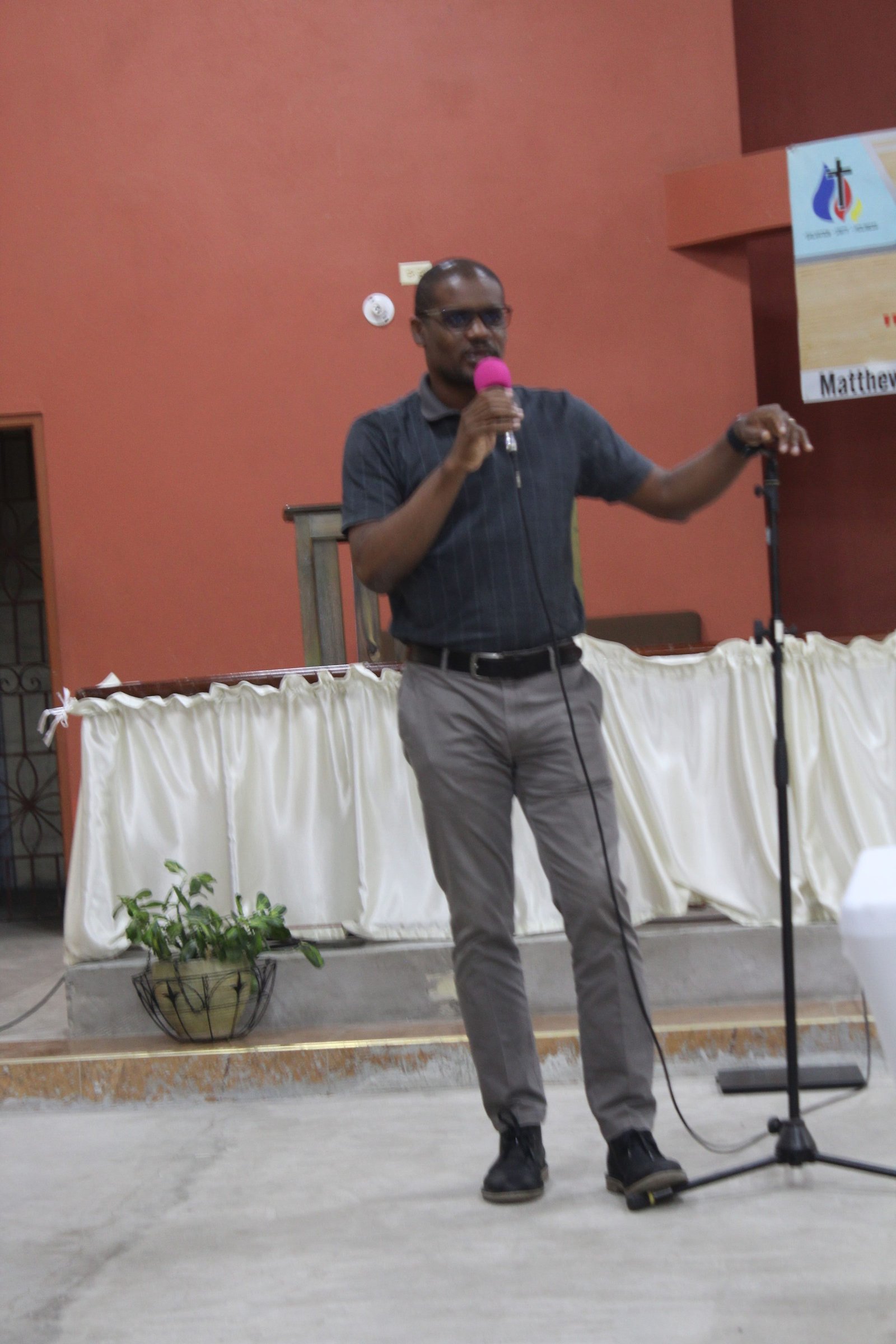 A man speaking into a microphone at a podium, wearing glasses and a short-sleeved shirt, with a seated audience in a room with a red wall.