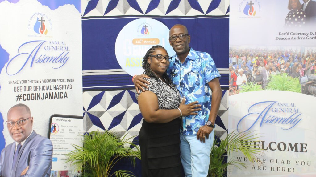 A couple posing together at an event booth with banners for the Annual General Assembly. The woman is wearing a black dress and the man is in a blue patterned shirt.