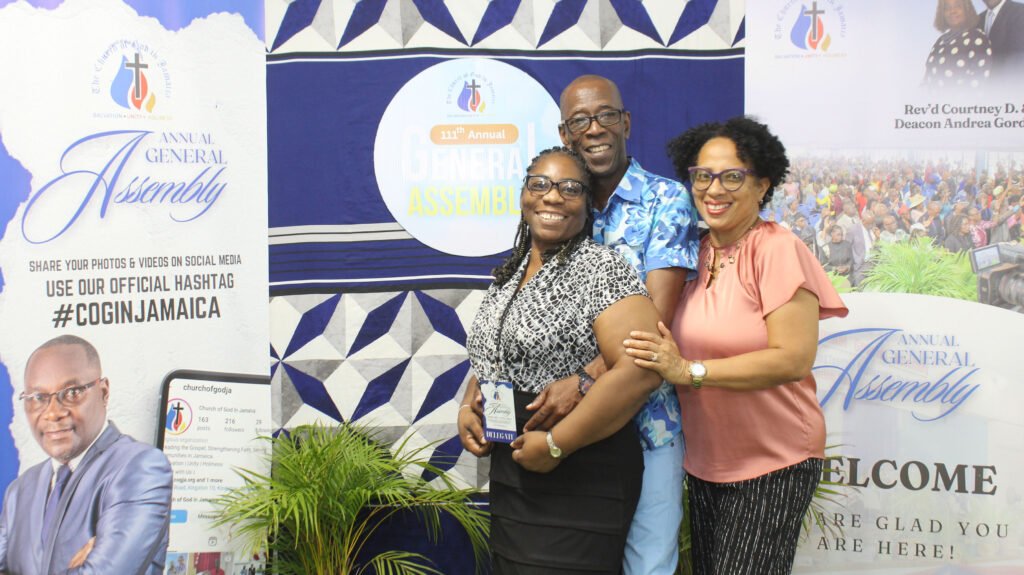 Three smiling individuals posing together in front of a decorative backdrop promoting the 'Annual General Assembly'. The backdrop features logos and information about the event, including hashtags for social media.