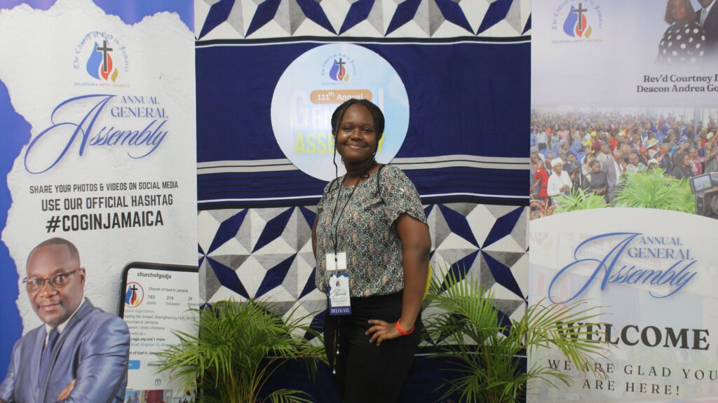 Woman posing at an event with banners displaying 'Annual General Assembly' and social media hashtags in the background.
