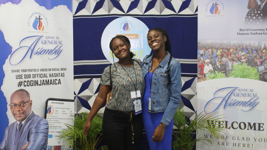 Two women posing together at a church event, with a backdrop featuring the Annual General Assembly branding and information.