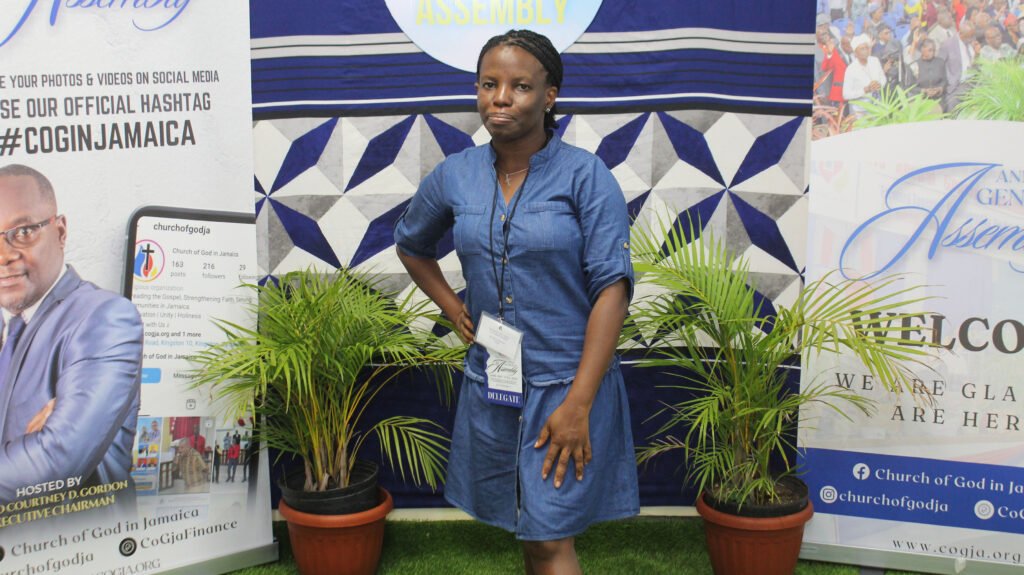 A woman stands in front of a backdrop featuring plants and promotional banners for the Church of God in Jamaica, displaying a welcoming message.