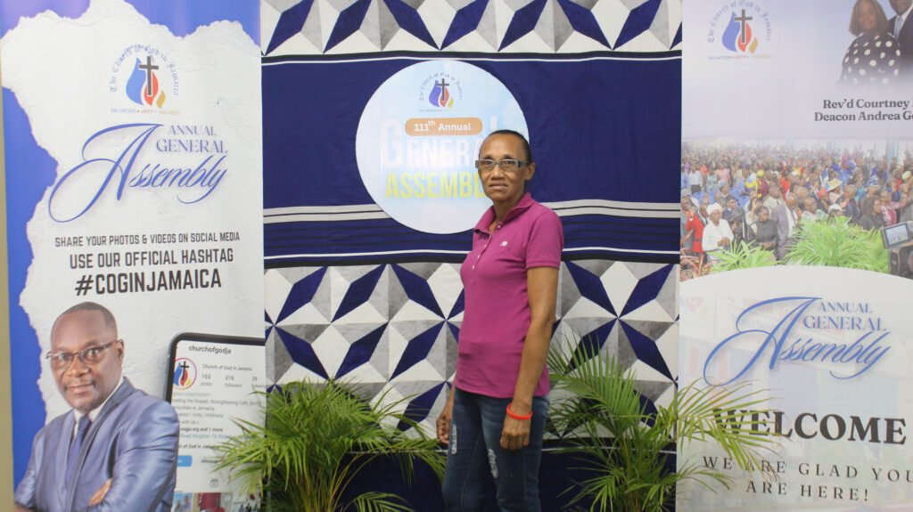 A woman in a pink shirt stands beside a backdrop promoting the Annual General Assembly of a church organization, featuring logos and hashtags for social media sharing.