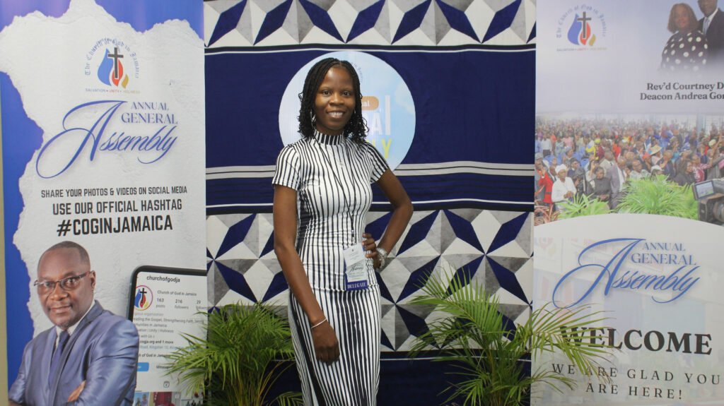 A woman in a striped dress stands smiling at an event surrounded by posters announcing the Annual General Assembly. The background features tropical plants and decorative fabric.