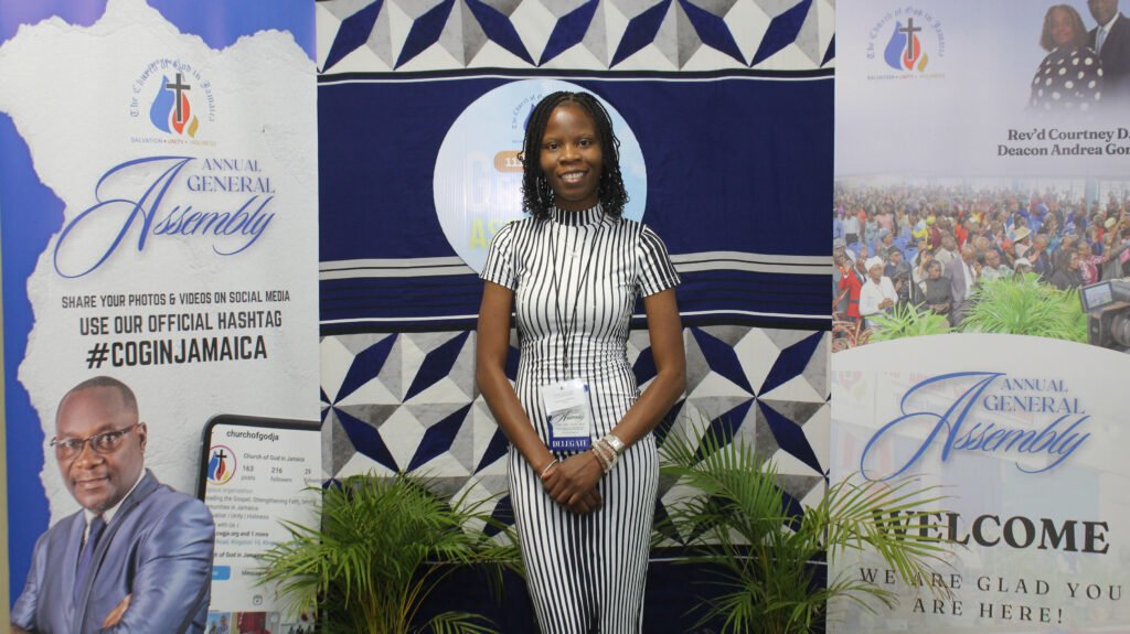 A woman standing in front of banners at the Annual General Assembly event, wearing a striped dress and holding a name badge.