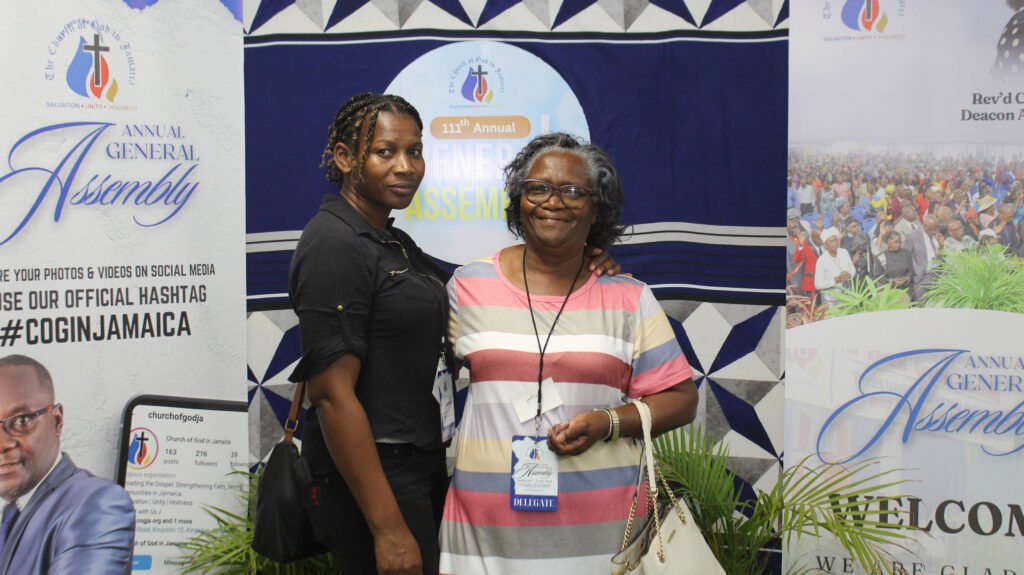 Two women posing together at an event, with banners and decorations in the background promoting the Annual General Assembly.