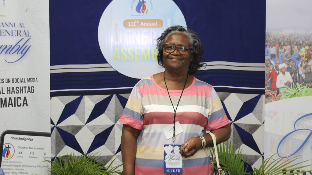 A woman smiles while standing in front of a banner for the 111th Annual General Assembly. She wears a striped shirt and a delegate badge.
