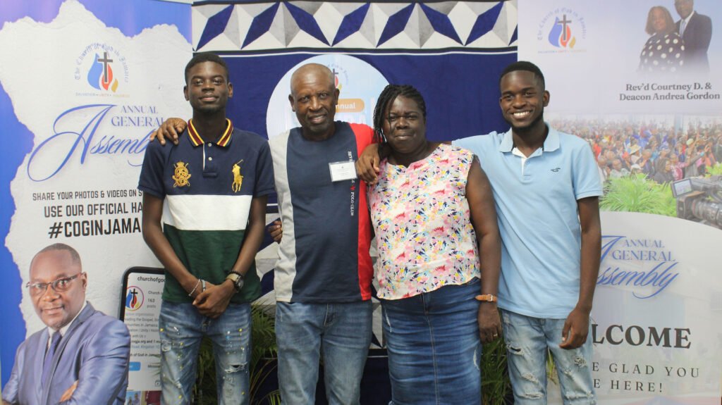 A group of four individuals standing together in front of a backdrop featuring promotional materials for an annual general assembly event.
