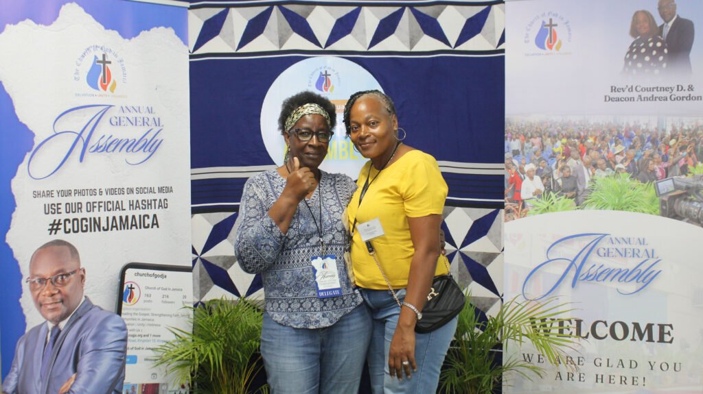 Two women posing together at an event, smiling at the camera. They are in front of a backdrop with banners announcing an Annual General Assembly.