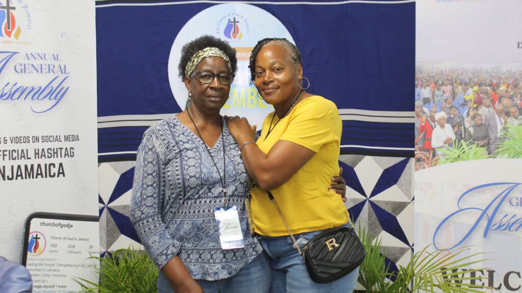 Two women smiling for a photo at a gathering, standing in front of a banner for the Annual General Assembly. One woman wears glasses and a patterned shirt, while the other wears a yellow top. The background features decorative elements and promotional materials for the event.