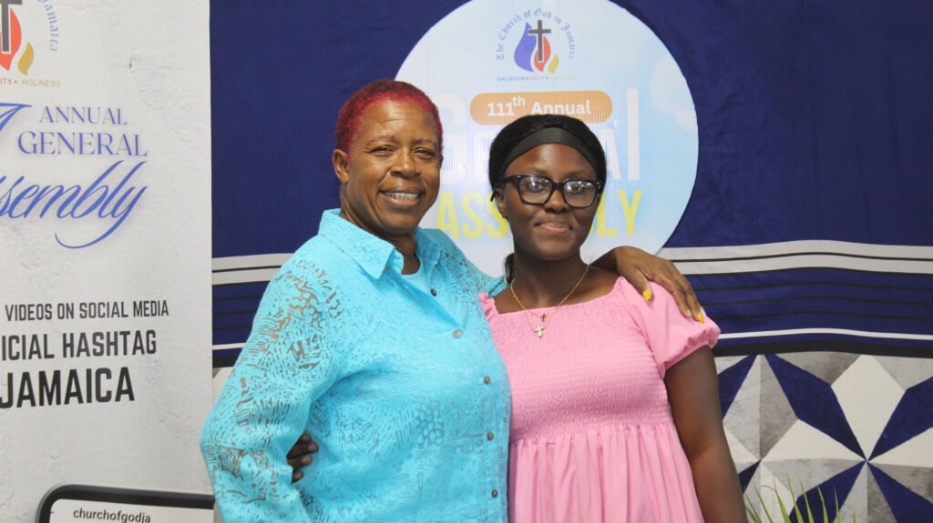 A woman with red hair and a blue shirt stands next to a young girl wearing glasses and a pink dress, both smiling in front of a backdrop that mentions an annual general assembly.