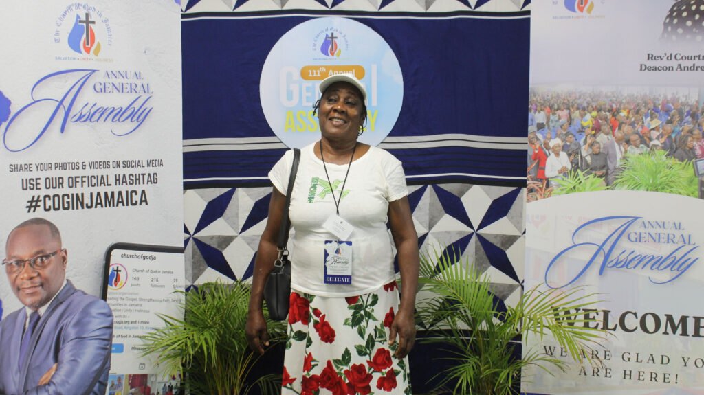A woman smiling and standing in front of a backdrop for the Annual General Assembly event, featuring banners with the event's logo and hashtags. She is wearing a white shirt and a floral skirt.