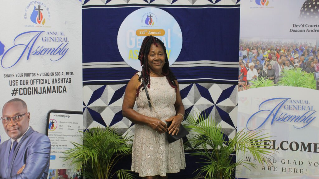 A woman stands smiling at an event, dressed in a light-colored dress, in front of banners for the Annual General Assembly, featuring logos and an encouraging message about sharing on social media.