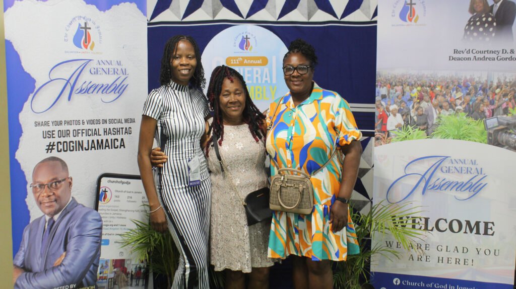 Three women posing together at an event, with banners in the background displaying the text 'Annual General Assembly' and the Church of God in Jamaica logo.